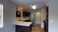 This single-floor residence showcases a thoughtfully designed interior that blends modern aesthetics with functional living. The kitchen serves as a central highlight, featuring sleek dark cabinetry that contrasts beautifully with light-toned countertops. A peninsula with an integrated double sink adds both workspace and a natural division between the kitchen and adjoining areas. Stainless steel appliances, including a full-size refrigerator and a black gas range, lend a contemporary edge to the space. Recessed and flush-mounted ceiling lighting ensures the kitchen remains bright and well-lit. Warm wood-look flooring flows throughout the space, adding a welcoming and cohesive feel underfoot. An arched architectural detail is visible in the background, providing a subtle decorative touch that softens the overall design. The neutral grey wall tones complement the dark cabinetry and light countertops, creating a balanced and inviting ambiance. The layout is efficient and open, allowing for ease of movement. Overall, the space presents a clean, modern interior suitable for comfortable everyday living.