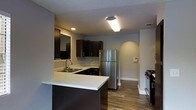 This single-floor residence showcases a thoughtfully designed interior that blends modern aesthetics with functional living. The kitchen serves as a central highlight, featuring sleek dark cabinetry that contrasts beautifully with light-toned countertops. A peninsula with an integrated double sink adds both workspace and a natural division between the kitchen and adjoining areas. Stainless steel appliances, including a full-size refrigerator and a black gas range, lend a contemporary edge to the space. Recessed and flush-mounted ceiling lighting ensures the kitchen remains bright and well-lit. Warm wood-look flooring flows throughout the space, adding a welcoming and cohesive feel underfoot. An arched architectural detail is visible in the background, providing a subtle decorative touch that softens the overall design. The neutral grey wall tones complement the dark cabinetry and light countertops, creating a balanced and inviting ambiance. The layout is efficient and open, allowing for ease of movement. Overall, the space presents a clean, modern interior suitable for comfortable everyday living.