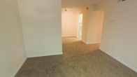 This single-floor residence offers a straightforward and functional layout designed for comfortable everyday living. The main living area is finished with wall-to-wall carpeting in a warm neutral tone, providing a soft underfoot feel throughout. Smooth, light-colored walls create a clean and airy backdrop, complemented by white baseboards that define each space crisply. A ceiling-mounted light fixture provides overhead illumination in the living area, contributing to a well-lit interior. The open layout allows the living space to flow naturally toward an adjacent area, which connects seamlessly to the kitchen. The kitchen is visible through a wide entryway and features white cabinetry and appliances, maintaining the consistent neutral palette found throughout the residence. A hallway branching off the main living space provides access to additional rooms, suggesting a well-organized floor plan. HVAC venting is integrated into the ceiling, ensuring climate control throughout. Overall, the space presents a clean, move-in-ready environment with a simple and cohesive aesthetic that offers a versatile foundation for personalization.