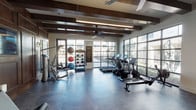 Located at 6107 Yellow Rock Trail in Dallas, TX, this single-floor fitness center combines refined architectural design with a fully equipped workout environment. The space is defined by its striking exposed dark wood ceiling beams, which lend a warm, rustic character that contrasts elegantly with the sleek, polished concrete flooring throughout. Oversized steel-framed glass walls and garage-style doors line the perimeter, flooding the interior with abundant natural light while offering open views of the surrounding landscaped exterior.

The cardio zone is well-stocked with a row of treadmills, elliptical trainers, and rowing machines, all positioned to take advantage of the expansive window views. A dedicated strength training area includes a multi-functional cable machine, a dual-arm weight machine, an adjustable bench, and a full dumbbell rack accompanied by a wall mirror. Fitness accessories are neatly organized on a dedicated rack and include medicine balls, kettlebells, stability balls, and TRX suspension trainers.

Rich dark wood wall paneling anchors one end of the space, adding a refined, club-like ambiance. Ceiling fans and pendant lighting provide practical comfort and visibility. Built-in cabinetry offers additional storage and utility, completing this thoughtfully designed, upscale fitness facility.