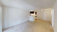 This single-story unit offers a practical and straightforward layout across several distinct spaces. The living area is carpeted in a neutral tone and features a textured ceiling, providing a clean, open canvas. A sliding glass door with vertical blinds allows natural light to fill the room and provides access to an outdoor area. The kitchen is designed in a galley configuration, featuring dark espresso-toned cabinetry on both sides, light countertops, a coil-top electric range, a full-size refrigerator, and a stainless steel sink. Track lighting overhead brightens the workspace efficiently. A pass-through or peninsula connects the kitchen to the main living space. The bedroom is a well-proportioned, carpeted room with a ceiling fan and light fixture, a window for natural light, and a closet. The bathroom is equipped with a single-sink vanity topped with a white countertop, dark wood cabinetry below, a large mirror, a standard toilet, and a full-size bathtub with a tiled surround and a decorative shower curtain. Tile flooring completes the bathroom.