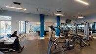 This single-floor fitness and recreation facility combines a comprehensive workout space with a social lounge area, unified by a contemporary design aesthetic. Bold blue structural columns punctuate the open layout, contrasting against light gray walls and durable hardwood-style flooring that transitions to rubberized gym matting in the exercise zones.

The fitness area is generously proportioned and well-equipped, featuring multiple treadmills, elliptical machines, cable weight systems by Cybex, a full dumbbell rack with adjustable benches, a rowing machine, and various resistance training equipment. Floor-to-ceiling windows along one wall offer direct views of an outdoor pool area surrounded by palm trees, providing both natural light and visual connection to the exterior amenity space. Large mirrors line the walls of the weight room, enhancing the sense of space while mounted flat-screen televisions offer entertainment during workouts. Linear LED lighting runs across the ceiling, ensuring consistent illumination throughout.

Adjacent to the gym, a lounge area features a full-size pool table with teal felt, complemented by a modern multi-ring pendant light fixture and colorful wall art. The open-concept design allows the two zones to flow seamlessly together, creating a well-rounded amenity space suited for both fitness and leisure.