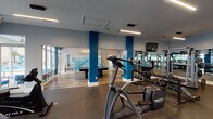 This single-floor fitness and recreation facility combines a comprehensive workout space with a social lounge area, unified by a contemporary design aesthetic. Bold blue structural columns punctuate the open layout, contrasting against light gray walls and durable hardwood-style flooring that transitions to rubberized gym matting in the exercise zones.

The fitness area is generously proportioned and well-equipped, featuring multiple treadmills, elliptical machines, cable weight systems by Cybex, a full dumbbell rack with adjustable benches, a rowing machine, and various resistance training equipment. Floor-to-ceiling windows along one wall offer direct views of an outdoor pool area surrounded by palm trees, providing both natural light and visual connection to the exterior amenity space. Large mirrors line the walls of the weight room, enhancing the sense of space while mounted flat-screen televisions offer entertainment during workouts. Linear LED lighting runs across the ceiling, ensuring consistent illumination throughout.

Adjacent to the gym, a lounge area features a full-size pool table with teal felt, complemented by a modern multi-ring pendant light fixture and colorful wall art. The open-concept design allows the two zones to flow seamlessly together, creating a well-rounded amenity space suited for both fitness and leisure.