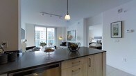 This contemporary single-floor residence showcases a thoughtfully designed open-concept layout that seamlessly connects the kitchen, living, and sleeping areas. The kitchen is a standout feature, centered around a spacious island with a dark stone countertop, light wood cabinetry, and gold-toned hardware. A pendant light hangs above the island, while a track lighting system illuminates the clean white ceiling above. Stainless steel appliances, including a built-in dishwasher, add a polished, modern touch. The living area flows naturally from the kitchen, bathed in natural light pouring through large floor-to-ceiling windows that frame impressive views of the surrounding cityscape. The space is furnished with a contemporary sofa and accented by a stylish floor lamp, creating a warm and inviting atmosphere. A framed artwork piece adds a creative focal point to the living area. Just off the main living space, the bedroom is visible through an open doorway, offering a neatly arranged sleeping area with a upholstered headboard. The overall aesthetic throughout is clean, modern, and cohesive, with a neutral palette of whites, grays, and warm wood tones creating a bright and sophisticated ambiance.