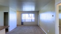 This single-floor space features an open living area finished with wall-to-wall carpeting that lends a warm, comfortable feel underfoot. The room is defined by a large picture window centered on the front wall, framed by sheer gray curtains that allow natural light to filter through while offering a view of the outdoors. The textured popcorn ceiling adds a classic architectural detail and contributes to the room's sense of height. Neutral-toned walls complement the overall palette, creating a clean and understated aesthetic throughout. A wall-mounted heating unit is positioned along one wall, providing a practical, space-saving climate control solution. A doorway to one side leads to an additional area of the unit, suggesting a straightforward and functional layout. White baseboards line the perimeter of the room, adding a crisp finishing detail against the carpeted floor. The overall ambiance is simple and utilitarian, offering a blank canvas well-suited for a variety of furnishing arrangements and personal customization.