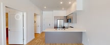 This contemporary 540-square-foot, one-bedroom, one-bathroom unit is located at 455 W 500 S in Salt Lake City, UT. The open-concept layout seamlessly connects the kitchen and living room, maximizing the efficient use of space. The kitchen is well-appointed with modern taupe flat-front cabinetry, stainless steel appliances including a refrigerator, gas range, over-the-range microwave, and dishwasher, complemented by a sleek white countertop peninsula with an integrated sink. Light hardwood flooring flows consistently throughout the entire unit, enhancing the bright, clean aesthetic. The living area features large floor-to-ceiling sliding glass doors and oversized windows that flood the space with natural light and offer impressive views of the Salt Lake City skyline and surrounding Wasatch Mountains. A ceiling fan adds comfort to the airy living space. The 150-square-foot bedroom includes a large window with additional city views. The bathroom features a full tub/shower combo, a vanity with taupe cabinetry and white countertop, and a large mirror. Clean white walls and recessed lighting throughout contribute to the unit's modern, minimalist ambiance.