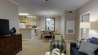 This single-floor residence features an open-concept layout that seamlessly connects the kitchen, dining area, and living room, creating a cohesive and functional living environment. The kitchen is outfitted with light wood cabinetry, white appliances including a refrigerator, microwave, and range, and a peninsula countertop that provides additional workspace and casual seating. Neutral beige carpeting extends throughout the main living areas, complementing the warm, taupe-toned walls. Crown molding runs along the ceiling perimeter, adding a refined architectural detail throughout the space. The dining area is anchored by a round white table surrounded by upholstered chairs in a classic style, set beneath a multi-bulb pendant chandelier that provides focused ambient lighting. A large framed botanical artwork serves as a striking focal point on the dining room wall. The living area is furnished with patterned accent chairs in blue, green, and gold tones, contributing color and visual interest. A ceiling fan is visible overhead, and HVAC vents are integrated into the walls, ensuring climate comfort. Overall, the space presents a clean, traditionally styled interior with consistent finishes throughout.