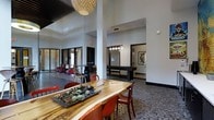 This single-floor space makes a strong visual impression through its dramatic double-height ceilings and an open, airy layout that encourages both social interaction and relaxed gathering. The main area centers around a striking live-edge wooden communal table paired with bold red chairs, anchored beneath a sculptural woven pendant light. A decorative succulent centerpiece adds a natural, organic touch to the otherwise contemporary setting.

The patterned hexagonal tile flooring extends throughout, providing a unifying design element that connects distinct zones within the space. Structural columns punctuate the open floor plan while contributing to the architectural character of the room. Natural light filters in through large entry doors and surrounding windows, keeping the interior bright and welcoming.

Toward the rear, a dedicated game room is visible, featuring a billiards table illuminated by a row of pendant lights, with framed artwork adorning the walls. A separate seating nook near the entrance offers a more intimate setting with additional chairs and tables. The overall ambiance blends industrial and eclectic design sensibilities, creating a versatile, communal environment suitable for a variety of activities.