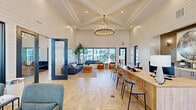 Located at 3755 E McKinney St in Denton, Texas, this single-floor community clubhouse blends modern farmhouse design with contemporary amenities across a generously sized open-plan layout. The space is defined by soaring vaulted and coffered ceilings, white shiplap walls, and light-toned herringbone wood flooring that flows throughout, creating a bright and cohesive aesthetic.

The welcoming lobby and reception area features a sleek wood bar-style counter with stylish seating, ideal for leasing or concierge functions. A statement ring chandelier anchors the entry, while large black-framed windows and glass-paned doors flood the interior with natural light and offer views of the surrounding exterior.

Adjacent lounge areas provide comfortable seating arrangements, including a casual sitting nook with a blue sofa, natural wood coffee table, and curated artwork on shiplap walls. A more expansive lounge features exposed dark wood ceiling beams, a bold geometric chandelier, and a large-screen TV, offering a relaxed media and social setting.

The recreation area centers around a designer billiards table beneath exposed beams, complemented by a full kitchenette bar equipped with a stainless steel refrigerator, counter seating, and vibrant accent wall murals. Throughout, abundant windows frame outdoor views, enhancing the open, airy ambiance of this well-appointed community gathering space.