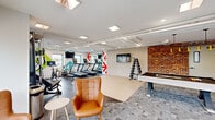 Located at 122 Bodman Place in Red Bank, NJ, this Colony House amenity space seamlessly combines a fully equipped fitness center with a vibrant social lounge. The open-plan layout is thoughtfully designed to accommodate both active and leisure activities within a single, cohesive environment.

The fitness zone features multiple treadmills, elliptical machines, a stationary bike, a rowing machine, a cable weight machine, and a dumbbell rack — all arranged for comfortable, open use. Bold graphic wall art with colorful arrow motifs energizes the workout area, while wall-mounted TVs and large windows providing views to the outdoors keep the space bright and engaging.

Transitioning into the lounge and game area, the space takes on a warmer, more relaxed character. A full-size pool table anchors this zone, complemented by plush seating including a navy velvet sofa and cognac leather accent chairs. Exposed brick walls, a striking living green wall, slatted wood paneling, and geometric gold pendant lights add rich texture and visual depth. The Colony House branding is subtly incorporated into the wall décor.

A dedicated open floor area with hardwood-style flooring provides flexibility for stretching or group activities. The polished concrete and carpet tile flooring throughout, exposed ceiling pipes, and recessed panel lighting contribute to a contemporary, industrial-chic aesthetic throughout the space.