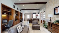 This single-story space showcases a thoughtfully designed open floor plan that seamlessly connects multiple functional zones. Exposed dark wood ceiling beams add architectural character throughout, complementing the warm, neutral color palette and earthy stone tile flooring that flows consistently across the entire space.

The main living area features comfortable leather seating arranged around a patterned area rug, while a striking natural limestone accent wall serves as a focal point, complete with a mounted flat-screen television and wooden credenza. Decorative metal star accents contribute a regional, rustic charm to the walls.

A built-in wooden workstation with dual computer stations, shelving, and cabinetry provides a dedicated workspace, finished in rich dark wood tones that echo throughout the design. Adjacent to this area, a round dining table with upholstered chairs offers an informal gathering spot.

The kitchenette features dark granite countertops with a stone-faced peninsula, dark cherry cabinetry, stainless steel appliances, and pendant lighting overhead. Leather bar stools line the counter, creating a casual seating option.

Elegant wood-and-glass French doors with transom windows flood the interior with natural light while providing access to an outdoor covered patio, enhancing the overall sense of openness and connectivity throughout the space.