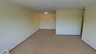 This single-floor space offers a straightforward and functional layout designed for comfortable everyday living. The interior features wall-to-wall carpet in a warm beige tone that extends throughout the open living area, contributing to a soft and understated aesthetic. The walls are finished in a neutral off-white palette, creating a clean and versatile backdrop that allows for a variety of furnishing styles. A flush-mount ceiling light provides central illumination to the main living area, keeping the design simple and unobtrusive. The layout includes an open-plan arrangement with a partial dividing wall that defines distinct zones within the space, offering a sense of separation without fully enclosing the areas. An intercom or thermostat panel is visible on the wall, indicating standard residential utilities are in place. The low-profile ceilings and neutral finishes give the space a modest, practical character. Overall, this is a clean, move-in ready environment that provides a blank canvas for personalization, suitable for a range of occupants looking for a no-frills, functional living space on a single level.