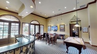 This single-floor community space is designed with a warm, lodge-inspired aesthetic that blends rustic charm with comfortable functionality. The entryway welcomes visitors with a spacious lounge area featuring plush sofas, upholstered armchairs, and decorative area rugs laid over tile flooring, all beneath soaring ceilings accented with rich dark wood crown molding. Arched French doors with transom windows line the walls, flooding the interior with natural light and offering views of an outdoor pool and courtyard area.

The game and activity room is a central highlight, furnished with a pool table, a shuffleboard table, and multiple dining-height game tables surrounded by leather-upholstered chairs. A wall-mounted flat-screen television, track lighting, and pendant fixtures add both practicality and ambiance. Decorative framed artwork, including equestrian-themed pieces, adds to the refined character of the space.

The fully equipped kitchen features dark wood cabinetry, granite countertops, a mosaic tile backsplash, and stainless steel appliances including a refrigerator, range, and microwave. Large format tile flooring runs throughout the kitchen and entry areas. Throughout the space, wrought iron decorative accents, recessed lighting, and warm neutral wall tones contribute to a cohesive, inviting atmosphere suited for gatherings and events.