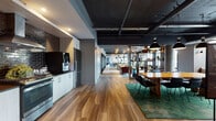 This expansive single-floor amenity space showcases a compelling industrial-modern aesthetic throughout. Exposed ductwork, concrete ceilings, and structural columns anchor the design, while warm hardwood flooring runs continuously through the main areas, creating visual cohesion across the layout.

The open-plan game and lounge area serves as the centerpiece, featuring a full-size pool table beneath oversized pendant lights, high-top seating, and generous wall art with a graphic typographic mural. Large windows line the perimeter, drawing in natural light and offering outdoor views.

A dedicated lounge room is finished in rich navy blue, furnished with plush sectional sofas, glass coffee tables, and framed artwork — providing an inviting retreat for relaxed gatherings.

The fully equipped kitchen includes stainless steel appliances, a gas range, a metallic brick tile backsplash, white cabinetry, dark countertops, and a smart beverage station. An adjacent dining area features a substantial wooden communal table.

A flexible activity zone with patterned carpet tile houses a children's play area and glass-partitioned private rooms. Near the entrance, bar-height counter seating along floor-to-ceiling windows creates an ideal spot for individual work or informal meetings.