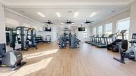 The Yardly Elm Fitness Studio at 4005 N Elm St in Denton, Texas is a thoughtfully designed, single-floor exercise facility offering a comprehensive workout experience. The space features light wood-tone flooring throughout, crisp white walls, and tray ceilings accented with large ceiling fans that maintain airflow across the generous open floor plan.

Natural light pours in through an abundance of large windows lining multiple walls, providing views of the surrounding community and outdoor amenity areas. The main workout area houses a full row of treadmills along one windowed wall, complemented by a multi-station cable and strength machine system positioned centrally. A dedicated free weights section includes a complete dumbbell rack, weight benches, and a squat/power rack, all positioned in front of full-length mirrors.

A separate cardio alcove features smart stationary bikes and a rowing machine, bathed in natural light from floor-to-ceiling windows overlooking the outdoor pool area. An open functional training zone provides ample floor space for dynamic workouts, equipped with medicine balls, kettlebells, resistance tools, and yoga mats on a dedicated rack. Wall-mounted televisions are strategically placed throughout, and a water station is conveniently located near the glass entry doors. The studio offers a bright, modern, and motivating environment suited for a wide range of fitness activities.