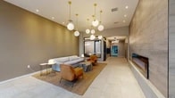Located at 4010 Lorain Ave in Cleveland's vibrant Ohio City neighborhood, this building offers a thoughtfully designed ground-floor experience that balances contemporary aesthetics with practical amenities. The welcoming lobby immediately draws attention with its striking cluster of globe pendant lights suspended from gold brass fixtures, casting a warm, inviting glow over the lounge area below. Comfortable seating — including a soft gray sofa, cognac leather armchairs, and natural stone side tables atop a woven jute rug — creates an elegant yet relaxed atmosphere. Light-toned tile flooring flows throughout, complementing the warm taupe wall palette. A glass-enclosed office or concierge space with black-framed partitions adds a polished, professional touch, while a bank of resident mailboxes lines the adjacent corridor. The hallway leads to elevators and an accessible restroom, with modern circular LED light fixtures overhead maintaining the building's refined design language. The entry vestibule features floor-to-ceiling glass walls and additional globe pendant lighting. Beyond the lobby, the building includes an expansive parking area with structural concrete columns, ample vehicle capacity, and large street-facing windows that allow natural light to filter in — providing convenient, covered parking for residents and visitors alike.