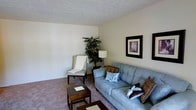 This single-floor residential space features a living room designed for comfort and everyday functionality. The living area is finished with wall-to-wall carpeting in a muted tone that complements the neutral, light-colored walls, creating a calm and cohesive atmosphere throughout. The textured ceiling adds a subtle architectural detail to the room.

The space is furnished with a light blue upholstered sofa accented with decorative throw pillows in complementary earth tones, paired with a classic white wingback accent chair draped with a casual throw blanket. A dark wood coffee table serves as a functional centerpiece, while a traditional-style table lamp provides warm, ambient lighting. A tall indoor plant in the corner introduces a natural element, softening the overall aesthetic.

Wall art in dark frames is displayed prominently, adding visual interest to the otherwise understated walls. The layout feels open and uncluttered, offering a straightforward, livable design. The overall ambiance is quiet and residential, with a practical arrangement suited for relaxed, everyday living.