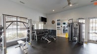 This single-floor fitness center is thoughtfully designed to support a comprehensive workout experience across multiple dedicated zones. The cardio area is lined with a row of Cybex treadmills and elliptical machines positioned along large windows that allow ample natural light to fill the space, complemented by wall-mounted flat-screen TVs for entertainment during workouts. Vaulted ceilings in this section enhance the open, airy feel.

The free weights area features a full rack of dumbbells alongside adjustable benches and a squat rack, all set against mirrored walls that amplify the sense of space. Ceiling fans and recessed lighting provide comfort throughout. A dedicated strength training zone houses multiple Cybex cable and resistance machines, with French doors opening to the exterior for added ventilation.

An accent wall in deep teal, adorned with a "Community" art piece, adds character and energy to the space. The connecting entryway is finished with warm wood-look flooring and a bold teal-and-white striped accent wall, featuring a built-in hydration station for convenience. Rubber flooring throughout the workout areas ensures safety and durability. Together, these zones create a functional, well-appointed fitness facility suited for a wide range of training needs.