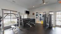 This single-floor fitness center is thoughtfully designed to support a comprehensive workout experience across multiple dedicated zones. The cardio area is lined with a row of Cybex treadmills and elliptical machines positioned along large windows that allow ample natural light to fill the space, complemented by wall-mounted flat-screen TVs for entertainment during workouts. Vaulted ceilings in this section enhance the open, airy feel.

The free weights area features a full rack of dumbbells alongside adjustable benches and a squat rack, all set against mirrored walls that amplify the sense of space. Ceiling fans and recessed lighting provide comfort throughout. A dedicated strength training zone houses multiple Cybex cable and resistance machines, with French doors opening to the exterior for added ventilation.

An accent wall in deep teal, adorned with a "Community" art piece, adds character and energy to the space. The connecting entryway is finished with warm wood-look flooring and a bold teal-and-white striped accent wall, featuring a built-in hydration station for convenience. Rubber flooring throughout the workout areas ensures safety and durability. Together, these zones create a functional, well-appointed fitness facility suited for a wide range of training needs.