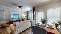 This single-story residence offers a thoughtfully designed open-concept layout connecting the living, dining, and kitchen areas into a cohesive, functional space. The living room is bright and welcoming, featuring crown molding, a ceiling fan, hardwood-style flooring, and tall windows dressed with floor-to-ceiling drapes that allow generous natural light. A wall-mounted television and a dedicated media console anchor one end of the room, while the dining area sits adjacent, making the space ideal for both everyday living and entertaining.

The kitchen is well-equipped with warm honey-toned cabinetry, white countertops, a subway tile backsplash, and black appliances including a range, microwave, refrigerator, and dishwasher. A peninsula with a built-in sink provides additional prep space and casual seating.

The bedroom features hardwood-style flooring, a ceiling fan, and a window with drape treatments that allow natural light. A walk-in closet with built-in shelving provides organized storage. The bathroom includes a dual-sink vanity with wood cabinetry, a large mirror, built-in shelving, and continued hardwood-style flooring, completing this cohesive and comfortable residence.