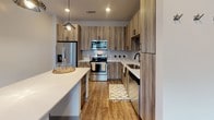 Located at 3705 SW Durham Dr in Durham, NC, this single-floor residence presents a contemporary design with a cohesive, modern aesthetic throughout. The open-concept living and kitchen area serves as the heart of the space, anchored by warm wood-look plank flooring that extends seamlessly across the main living zones. The kitchen is well-appointed with stainless steel appliances, including a refrigerator with ice dispenser, an electric range, and an over-the-range microwave. Flat-front cabinetry in a natural wood-tone finish reaches toward the ceiling, maximizing storage, while a white quartz-topped center island with pendant lighting overhead provides both prep space and casual dining potential. A subway tile backsplash adds a clean, polished accent. The bedroom features plush carpeting, a ceiling fan, and windows that invite natural light. The bathroom is notably spacious, with an extended double vanity topped in white quartz, wood-tone cabinetry with matte black hardware, a glass-enclosed shower with subway tile surround, and direct access to a walk-in closet with built-in shelving. Recessed lighting and a neutral color palette create a bright, cohesive ambiance throughout the unit.
