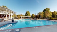 This expansive outdoor amenity space is designed for relaxation and recreation, centered around a large, resort-style swimming pool with crystal-clear blue water. The pool area features a classic brick-edged surround and is complemented by a pergola structure that provides shaded seating, making it ideal for warm-weather gatherings. Mature trees frame the perimeter, offering a natural backdrop and added privacy.

Adjacent to the pool, a generously sized concrete patio serves as an outdoor living area equipped with plush, navy blue upholstered lounge furniture, including a sectional sofa, chaise lounges, and a coffee table. The seating arrangement is positioned against a prominent textured retaining wall constructed of interlocking stone blocks, which adds architectural character and defines the space.

The patio also features dedicated grilling stations, including a built-in stainless steel grill and a ceramic kamado-style charcoal grill, both set on brick bases with granite countertops—ideal for outdoor cooking and entertaining. Decorative lamp posts provide ambient lighting throughout the area.

A separate hot tub is situated near the main building, which features a white-columned balcony, brick exterior, and an exterior staircase. The overall ambiance is polished, welcoming, and well-maintained.