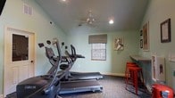 Located at 1175 Mathis Ferry Rd in Mt. Pleasant, South Carolina, this single-floor fitness facility offers a well-equipped and functional space for residents and users seeking a convenient workout environment. The room features vaulted ceilings accented by a ceiling fan and recessed lighting, which together create an open and airy atmosphere. The walls are finished in a soft sage green, lending the space a calm and motivating ambiance. A window with a decorative valance allows natural light to filter into the room, brightening the interior. The fitness area is outfitted with SportsArt cardio machines, including treadmills and elliptical trainers, positioned to allow comfortable use. A wall-mounted "Gym" sign adds a touch of personality and character to the space. Along one side, a countertop surface paired with red metal bar stools provides a convenient spot for users to set down belongings or take a brief rest. The carpeted flooring adds a degree of comfort underfoot while helping to define the space as a dedicated exercise area. Overall, the facility is thoughtfully designed to serve as a practical and welcoming fitness destination within the Mt. Pleasant community.