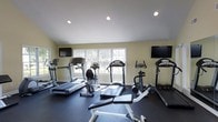 This dedicated fitness space is thoughtfully designed to support a comprehensive workout experience within a single-level layout. The room features smooth white ceilings with recessed lighting that provides even, consistent illumination throughout the space. Large multi-pane windows along one wall invite an abundance of natural light, creating a bright and energizing atmosphere while offering views of the surrounding greenery outdoors.

The flooring consists of dark, durable rubber matting — ideal for high-impact exercise — which contrasts cleanly with the light yellow-toned walls. The space is outfitted with a variety of cardio equipment, including multiple treadmills, a stationary recumbent bike, and an elliptical machine, offering users a range of workout options. A weight bench with barbell equipment is also present for strength training.

Two flat-screen televisions are mounted on opposite walls, ensuring visibility from multiple workout stations and adding an element of entertainment during exercise sessions. The layout is organized and spacious, allowing users to move freely between equipment. Overall, the space strikes a balance between functionality and comfort, making it a practical and well-appointed fitness facility.