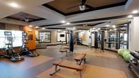 This fitness center offers a spacious, single-level layout designed to accommodate a comprehensive workout experience. The flooring features a two-tone design, combining neutral and warm terracotta-toned rubberized surfaces that delineate workout zones while providing durability and safety underfoot. The ceiling is a notable architectural feature, with a tray-style design accented by bold dark trim that adds visual depth and dimension to the space. A ceiling fan is centrally mounted, promoting air circulation throughout.

The facility is well-lit with recessed lighting positioned throughout, ensuring consistent illumination across all workout areas. Large mirrors are mounted along the walls, enhancing the sense of openness and allowing users to monitor their form during exercise. The walls are painted in a combination of warm orange and neutral tones, contributing to an energetic yet approachable atmosphere.

Equipment includes multiple cable and weight machines, adjustable benches, and cardio equipment such as treadmills, visible both directly and reflected in the mirrors. The open central floor space provides ample room for stretching or functional training. Overall, the space is clean, organized, and thoughtfully arranged to support a variety of fitness activities.