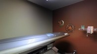 This single-floor tanning salon offers a compact yet well-appointed space designed to provide a comfortable and relaxing experience. The room is defined by a rich, warm brown accent wall that sets an inviting tone and complements the sun-inspired aesthetic throughout. A professional-grade tanning bed serves as the centerpiece of the space, equipped with modern lighting technology and a sleek, streamlined design built for client comfort and effective use. The walls are adorned with decorative sun and moon metal wall art, adding a thematic and visually engaging touch that reinforces the sun-soaked ambiance of the environment. Neutral tones on the remaining walls balance the deeper accent color, keeping the space feeling open despite its intimate scale. The interior is purposefully arranged to maximize functionality while maintaining a welcoming atmosphere. The overall design reflects a thoughtful approach to creating a relaxing, sun-themed retreat, making it well-suited for a personal or boutique tanning studio setting. The warm color palette and decorative details work in harmony to evoke a sense of warmth and leisure from the moment one enters the space.