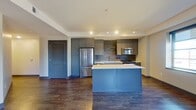 This well-appointed one-bedroom, one-bathroom unit offers a thoughtfully designed layout across a single floor, making efficient use of its comfortable footprint. The open-concept kitchen and living area serves as the centerpiece of the unit, highlighted by rich hardwood-style flooring, recessed and track lighting, and a bold blue kitchen island with a crisp white countertop. The kitchen is equipped with full stainless steel appliances, including a French door refrigerator, built-in microwave, and dishwasher, complemented by floor-to-ceiling grey cabinetry and a textured tile backsplash. Multiple windows in the living area allow natural light to fill the space while framing urban views. The bedroom features plush carpeting, a ceiling fan, and its own window with city views, providing a quiet retreat. A dedicated walk-in closet with built-in shelving and hanging rods offers ample storage. The bathroom impresses with a glass-enclosed walk-in shower tiled in classic white subway tile with accent detailing and a built-in bench. A connecting hallway with hardwood-style flooring ties the layout together cohesively.
