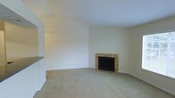 This single-floor space centers around a comfortable living area distinguished by vaulted ceilings that lend an open, airy quality to the room. The focal point is a built-in fireplace framed by warm-toned square ceramic tiles, set against a crisp white surround that extends to the ceiling, creating a striking architectural feature. The hearth area transitions from carpet to a tiled floor section, providing a practical and visually defined zone around the fireplace. Neutral beige carpeting covers the main floor area, contributing to a warm and understated atmosphere throughout. The walls are finished in a clean, off-white tone that reflects light and keeps the space feeling bright and open. A white interior door with brass hardware provides access to an adjacent area, adding to the functional layout. The overall design is simple and understated, with the vaulted ceiling and fireplace serving as the primary architectural highlights. The neutral palette and open feel make this space highly adaptable for a variety of furniture arrangements and interior styles.