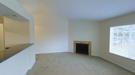 This single-floor space centers around a comfortable living area distinguished by vaulted ceilings that lend an open, airy quality to the room. The focal point is a built-in fireplace framed by warm-toned square ceramic tiles, set against a crisp white surround that extends to the ceiling, creating a striking architectural feature. The hearth area transitions from carpet to a tiled floor section, providing a practical and visually defined zone around the fireplace. Neutral beige carpeting covers the main floor area, contributing to a warm and understated atmosphere throughout. The walls are finished in a clean, off-white tone that reflects light and keeps the space feeling bright and open. A white interior door with brass hardware provides access to an adjacent area, adding to the functional layout. The overall design is simple and understated, with the vaulted ceiling and fireplace serving as the primary architectural highlights. The neutral palette and open feel make this space highly adaptable for a variety of furniture arrangements and interior styles.