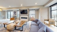 Located at 500 W Lake St in Minneapolis, this well-appointed community amenity space offers a thoughtfully designed layout across a single level. The open-concept living area serves as the heart of the space, featuring a sleek wall-mounted fireplace with a large flat-screen TV above, surrounded by contemporary furnishings set atop patterned area rugs and light wood-style flooring. Abundant natural light pours in through large windows and sliding glass doors that lead to two outdoor balconies, offering panoramic views of the Minneapolis skyline and surrounding neighborhoods. One rooftop balcony is outfitted with lounge seating, a central fire pit, and festoon lighting, creating an inviting outdoor gathering area. The kitchen is a standout feature, showcasing bold cobalt blue cabinetry, a striking marble-veined quartz countertop and backsplash, stainless steel appliances, and a spacious island with woven rattan barstools beneath globe pendant lights. The dining area accommodates groups at long communal high-top tables beneath sculptural pendant lighting, with city views as a backdrop. Vibrant curated artwork throughout adds personality, while textured wall treatments and recessed lighting lend the space a polished, modern ambiance ideal for gatherings and social events.