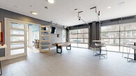 This thoughtfully designed single-floor amenity space offers a generous and versatile layout spanning a substantial footprint. The space is organized into distinct functional zones connected by a central hallway, each reflecting a cohesive modern aesthetic.

The kitchen and living room area serves as a social hub, featuring a U-shaped kitchen outfitted with dark granite countertops, gray shiplap backsplash, stainless steel appliances, and large-format tile flooring. Adjacent lounge areas display a striking teal-painted ceiling, exposed brick accent walls, and colorful artwork, creating a vibrant atmosphere. Comfortable seating arrangements and high-top tables make this zone ideal for gathering and relaxation.

A game and social room showcases industrial-style overhead garage doors that flood the space with natural light and views of the surrounding greenery. Light tile flooring, a shuffleboard table, wall-mounted television, and bar-height seating round out this lively entertainment area.

The dedicated exercise room is well-appointed with treadmills, ellipticals, free weights, and organized equipment storage. A bold teal ceiling, exposed brick columns, large windows, and ceiling fans contribute to an energizing workout environment. Carpeted flooring and ample natural light enhance the space's functionality and comfort throughout.