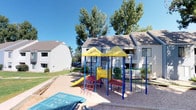 This shared outdoor common area serves as the central gathering space for the surrounding residential community. The grounds are well-maintained, featuring expansive stretches of lush green lawn interspersed with mature trees that provide ample shade throughout the property. Multiple communal charcoal BBQ grills are strategically positioned on concrete pads across the grounds, making outdoor cooking and entertaining convenient for residents.

A colorful children's playground is a notable amenity, equipped with slides, climbing structures, and play equipment set within a safety-surfaced area. The playground is surrounded by mature trees, offering a shaded and inviting environment for younger residents.

The buildings themselves feature stucco exteriors in neutral tones, with covered balconies and white railed porches at both ground and upper levels. Landscaping throughout the property combines gravel beds, rock borders, and planted shrubs alongside the green lawns, creating a varied and tidy aesthetic. Pathway lighting is provided by globe-style lamp posts positioned throughout the grounds.

Practical infrastructure, including HVAC units, is discreetly placed along building exteriors within gravel-bordered zones. The overall ambiance of the outdoor space is open, functional, and community-oriented, offering residents a variety of recreational and leisure options in a well-kept setting.