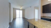 This single-floor space showcases a thoughtfully designed open-concept layout that seamlessly integrates the kitchen and living areas. The flooring throughout consists of rich, dark hardwood-style planks that contrast elegantly with the bright, neutral-toned walls and white trim, creating a clean and polished aesthetic.

The kitchen area features a striking granite countertop with warm golden and brown tones, adding a touch of sophistication to the space. The living area is anchored by a ceiling fan, which enhances both comfort and airflow, while a large sliding glass door floods the room with abundant natural light and provides direct access to a private balcony. The balcony offers views of the surrounding exterior, making it an appealing extension of the living space.

A doorway visible toward the rear of the unit leads to additional rooms, suggesting a functional layout with clear separation between living and private spaces. The overall ambiance is bright, modern, and move-in ready, with design choices that balance practicality with visual appeal. The space presents a well-maintained interior suited for comfortable everyday living.