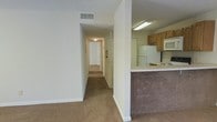 This single-story space is designed with a practical, straightforward layout. A central hallway extends toward the back of the unit, lined with neutral-toned carpet and white walls, connecting the living area to the additional rooms beyond. The hallway is well-lit by a ceiling fixture and features a fire extinguisher mounted on the wall, underscoring safety considerations built into the space.

Adjacent to the main living area, a kitchen is partially separated by a half-wall that doubles as a breakfast bar, offering a casual dining or workspace surface. The kitchen is equipped with light oak cabinetry, a white refrigerator, a built-in microwave, and a stovetop, providing all essential appliances in a compact yet functional arrangement. An overhead fluorescent light fixture illuminates the kitchen workspace effectively.

The flooring throughout the living and hallway areas consists of beige carpet, contributing to a warm, understated aesthetic. Ceiling-mounted HVAC vents are visible, ensuring climate control throughout the space. The overall interior presents a clean, neutral palette with white walls and trim, creating a versatile backdrop adaptable to a variety of furnishing styles.