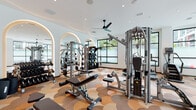 Located at 3850 S Bond Avenue in Portland's South Waterfront neighborhood, this well-appointed fitness center offers a comprehensive workout environment within a single, open-plan floor. The space is immediately distinguished by its eye-catching scallop-patterned carpet in warm earth tones of tan, brown, and terracotta, which extends throughout the entire facility.

The strength training area is generously equipped with multiple multi-station cable machines, adjustable benches, a full dumbbell rack, and functional training accessories. A striking row of illuminated arched mirrors lines one wall, visually expanding the space and providing excellent form-checking capability during workouts. Overhead, modern ceiling fans ensure comfortable air circulation throughout.

The cardio zone is positioned along a wall of large, black-framed windows that flood the space with natural light and offer views of the surrounding urban landscape and greenery. Equipment includes treadmills, elliptical trainers, and stationary bikes. A dedicated alcove with floor-to-ceiling windows provides an especially bright and open setting for cardio exercise.

Additional amenities include a wall-mounted television, a built-in beverage cooler, and storage for accessories, alongside curated wall art that lends the space a refined, contemporary aesthetic. Recessed lighting throughout ensures the facility is well-lit at all hours.