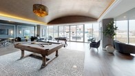 This expansive single-floor amenity lounge is defined by its striking architectural character and thoughtfully curated interior design. A distinctive curved barrel-vaulted ceiling with a richly textured, dark metallic finish serves as a focal point throughout the space, accented by warm amber LED cove lighting that creates an inviting glow. Light hardwood flooring flows continuously across the open-plan layout, unifying the various functional zones.

Floor-to-ceiling glass walls along one side of the lounge flood the interior with natural light while framing panoramic city views and providing access to an outdoor terrace. This glazed perimeter hosts intimate seating clusters with sculptural velvet armchairs, ideal for quiet conversation.

The lounge is organized into distinct areas: a cinema-style seating zone features curved, deep-toned velvet sectional sofas arranged around a wall-mounted television framed by classic wainscoting and black-and-white artwork. A games area is equipped with a rustic wood billiards table and a foosball table beneath an ornate tiered amber chandelier. A separate lounge section includes softer seating arrangements and a linear fireplace set into a paneled accent wall. The space balances upscale social function with relaxed elegance throughout.