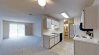 This single-floor residence offers a functional and straightforward layout designed for comfortable everyday living. The kitchen serves as a central hub of the home, featuring white cabinetry that provides generous storage space, a double-basin sink, a dishwasher, a refrigerator, and a range. A skylight-style ceiling fixture brightens the kitchen workspace, while a ceiling fan with a globe light adds additional illumination and airflow to the adjacent area. The kitchen flows openly into the carpeted living area, which benefits from natural light streaming in through a large sliding glass door dressed with vertical blinds — hinting at outdoor access. Light-toned walls and neutral-colored carpet throughout create a clean, understated aesthetic. A hallway visible from the kitchen leads to what appears to be a bathroom, with vanity lighting visible in the background, suggesting well-connected and accessible room arrangements. The light tile flooring in the kitchen transitions seamlessly to the soft carpeting of the living space, distinguishing functional zones while maintaining a cohesive overall feel. The space presents a practical, move-in-ready environment with a simple, uncluttered design.