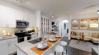 This single-story, 2-bedroom, 2-bathroom residence is designed with a modern aesthetic and an open-concept floor plan that promotes a seamless flow between living spaces. The kitchen serves as a central highlight, featuring sleek white cabinetry, stainless steel appliances — including a range and built-in microwave — and a striking granite-topped island with an undermount sink. The island doubles as a breakfast bar with white bar stools, making it ideal for casual dining or entertaining. A mosaic tile backsplash adds texture and visual interest to the cooking area. The kitchen opens directly into the dining and living areas, creating a cohesive, airy atmosphere. An arched architectural detail frames the hallway leading toward the bedrooms, adding a refined design touch. Dark hardwood-style flooring runs throughout the main living areas, complementing the crisp white walls and crown molding. Decorative wall art and warm accent colors contribute to an inviting ambiance. The overall layout is thoughtfully arranged to maximize functionality and comfort across both bedrooms and bathrooms, offering a well-appointed living experience.
