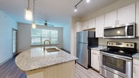 Located at 10018 Ingram Rd in San Antonio, TX, this one-bedroom, one-bathroom unit offers a well-designed, single-floor layout that maximizes functionality and comfort. The open-concept kitchen and living area serves as the heart of the home, anchored by a curved granite-topped island with an undermount sink and elegant pendant lighting. The kitchen is outfitted with stainless steel appliances, including a refrigerator, range, and built-in microwave, complemented by light wood cabinetry that extends to the ceiling for generous storage. Track lighting overhead adds a modern touch. Wood-style flooring flows throughout the main living areas, lending warmth and visual continuity. The living room benefits from large windows and a sliding glass door that flood the space with natural light, while a ceiling fan adds comfort. The spacious primary bedroom connects to a walk-in closet with wire shelving and an additional secondary closet for ample storage. The bathroom features tile flooring, a granite vanity with wood-toned cabinetry, and a full tub-shower combination. A dedicated laundry area houses a stacked washer and dryer unit, adding everyday convenience. The layout is efficient and thoughtfully organized throughout.