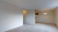 This single-floor space presents an open-concept layout that seamlessly connects the living area to the kitchen. The living space is finished with light neutral carpeting that extends throughout, contributing to a clean, understated aesthetic. Smooth white walls and a flat ceiling amplify the sense of openness, while a partial dividing wall subtly separates the kitchen from the main living area without fully enclosing it.

The kitchen is visible from the living space and features warm-toned wood cabinetry positioned above a white refrigerator, along with a dark countertop that provides functional contrast. A chandelier with multiple candle-style lights hangs at the transition between the kitchen and dining area, adding a decorative focal point to an otherwise straightforward layout.

The overall ambiance is bright and neutral, with the white walls and light flooring working together to maximize the sense of space. The open flow between living and kitchen areas makes the unit feel cohesive and functional, well-suited for everyday living.