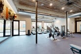 Located in Charlotte, NC, this fitness center offers a thoughtfully designed exercise space that blends industrial and rustic aesthetics seamlessly. The standout architectural feature is the reclaimed wood ceiling, which adds warmth and character, complemented by exposed metal ductwork and large ceiling fans that maintain airflow throughout the space. Structural steel columns punctuate the open floor plan, reinforcing the industrial character while preserving a spacious, airy feel.

Natural light flows generously through large, dark-framed windows and glass doors along multiple walls, creating a bright and energizing environment that opens to outdoor views. The flooring throughout is durable, blue-toned commercial carpet tile, well-suited for high-traffic fitness use.

The cardio zone features a row of treadmills and elliptical machines positioned along the windowed walls, while stationary and cycling bikes occupy the central area. A dedicated strength training section includes a multi-function cable machine, a power rack with bench and weight plates, and additional resistance equipment. An exposed brick accent wall with decorative industrial fan elements adds visual interest to the strength area, alongside floor-to-ceiling mirrors that enhance the sense of depth. The space is illuminated by recessed lighting, providing consistent, even coverage throughout.