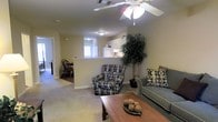 This single-floor residence features an open-concept design that seamlessly connects the living, dining, and kitchen areas. The living room is a comfortable and welcoming space, centered around a wood coffee table and furnished with a patterned accent chair and a grey sofa accented with coordinating throw pillows. A ceiling fan with a light fixture overhead provides both air circulation and ambient lighting. A large decorative potted tree adds a natural element to the corner of the room, enhancing the warm atmosphere.

The kitchen is visible beyond a breakfast bar counter that separates it from the living area. White cabinetry lines the walls, and standard appliances, including a refrigerator, are visible. The bar counter provides a casual dining or prep surface and keeps the space feeling open and connected.

Adjacent to the kitchen, a dining area accommodates a wooden table and chairs, bathed in natural light from a nearby window. A hallway visible from the living room suggests additional rooms beyond, maintaining a functional and organized flow throughout the space. Neutral wall tones and carpet flooring unify the overall aesthetic.