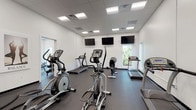 This single-level fitness center is thoughtfully designed to accommodate a full range of exercise needs within a clean, modern environment. The space is divided into distinct functional zones, each outfitted with professional-grade equipment. The cardio area features multiple treadmills, elliptical trainers, stationary bikes, and rowing machines arranged to allow comfortable spacing between users. Wall-mounted flat-screen televisions are positioned throughout this section, providing entertainment during workouts. Natural light filters in through several windows, offering views of outdoor greenery and contributing to an energizing atmosphere.

The strength training zone is equally well-appointed, housing a variety of cable machines, multi-station weight systems, free weight benches, and a dumbbell rack. Large wall mirrors span multiple sections, enhancing the sense of space while allowing users to monitor their form. A bank of lockers along one wall provides secure personal storage. The dark rubber flooring throughout offers both durability and safety underfoot. Recessed panel lighting in the drop-tile ceiling ensures the entire space is evenly and brightly illuminated. The crisp white walls and organized layout contribute to a functional, professional ambiance suited for serious training.