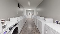 This shared laundry facility is designed with practicality and efficiency in mind, offering a generous number of commercial-grade washing machines and dryers arranged in parallel rows along both sides of the room. The elongated layout creates a clear central walkway, allowing easy access to all machines simultaneously. The white appliances stand out against the neutral gray walls and polished concrete flooring, giving the space a clean, utilitarian aesthetic.

Overhead fluorescent lighting ensures the room is well-lit throughout, while wall-mounted HVAC vents support adequate ventilation — an important feature in a high-use laundry environment. A dedicated utility area at one end of the facility includes a deep white utility sink paired with a speckled granite-style folding counter that runs along the wall, providing a practical surface for sorting and folding laundry. Instructional signage and a fire extinguisher are visible, reflecting attention to safety and user guidance.

The space is accessed through a standard door marked with laundry room signage, and the overall design prioritizes function and durability. The combination of ample machine capacity, folding space, and a well-maintained environment makes this a highly functional communal laundry facility.