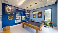 Located at 3300 Boutwell Rd in Lake Worth Beach, Florida, this sports-themed game room is a dynamic and well-appointed recreational space designed for entertainment and socializing. The room is defined by bold cobalt and steel-blue walls adorned with a curated gallery of sports artwork, including aerial stadium photography, Formula 1 racing prints, and classic sports imagery, creating an energetic and immersive atmosphere.

At the center of the space sits a handsome wood-framed pool table with a dark felt surface, positioned beneath a trio of oversized circular pendant lights that cast a warm, focused glow. High-gloss white tile flooring and a tray ceiling add a polished, contemporary feel. A large flat-screen TV mounted on the feature wall serves as a media focal point, complemented by a dartboard and a connect-four game mounted nearby for additional entertainment options.

Tall, floor-to-ceiling windows and a glass door flood the space with natural light and frame lush greenery and landscaping outside, seamlessly connecting the interior to the outdoors. A bistro-height table with leather bucket chairs provides casual seating for spectators. A glass partition wall offers a glimpse into an adjacent lounge area, enhancing the sense of openness. This thoughtfully designed game room delivers a compelling blend of sporty character and modern sophistication.