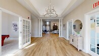 Located at 39 Buckland St in Manchester, CT, this single-floor community space offers a polished and welcoming atmosphere defined by classic architectural design and thoughtful interior styling. The entrance greets visitors with soaring tray ceilings, intricate crown molding, and a striking dual-tier chandelier that serves as a dramatic focal point. Natural light pours in through a transom-topped set of French doors, flanked by additional French-door side panels, giving the foyer an airy, open feel. Warm-toned hardwood flooring flows seamlessly throughout the space, complementing the neutral wall palette. White decorative columns frame the transition from the entrance into the expansive game room, where the refined aesthetic continues. The game room features a stone-surround fireplace with a wall-mounted television above, floor-to-ceiling drape-framed windows, and recessed lighting. Comfortable seating arrangements with contemporary furnishings in soft grays and navy blues create an inviting lounge environment. A distinctive arched wall niche with flanking columns and wooden shuttered details adds a unique architectural accent. A dining area with bold blue accent chairs rounds out the space, making it equally suitable for social gatherings or relaxed recreation.